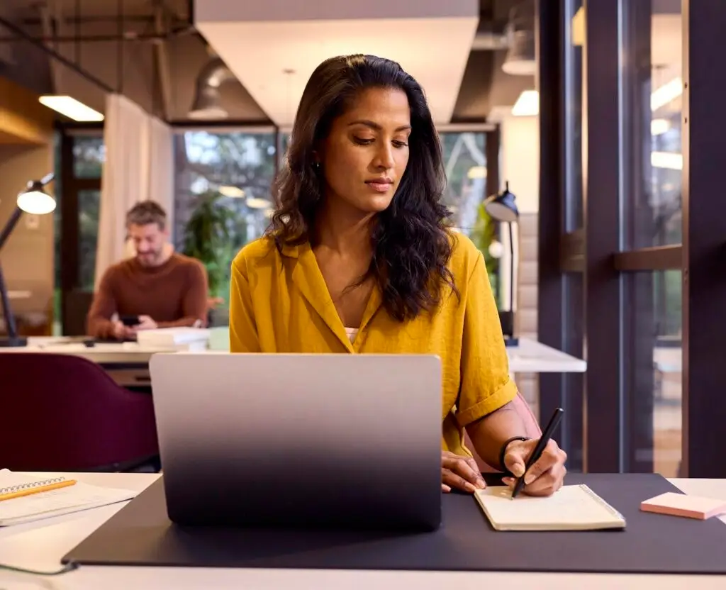 Mature Businesswoman Working On Laptop At Desk In Office Writing In Notebook