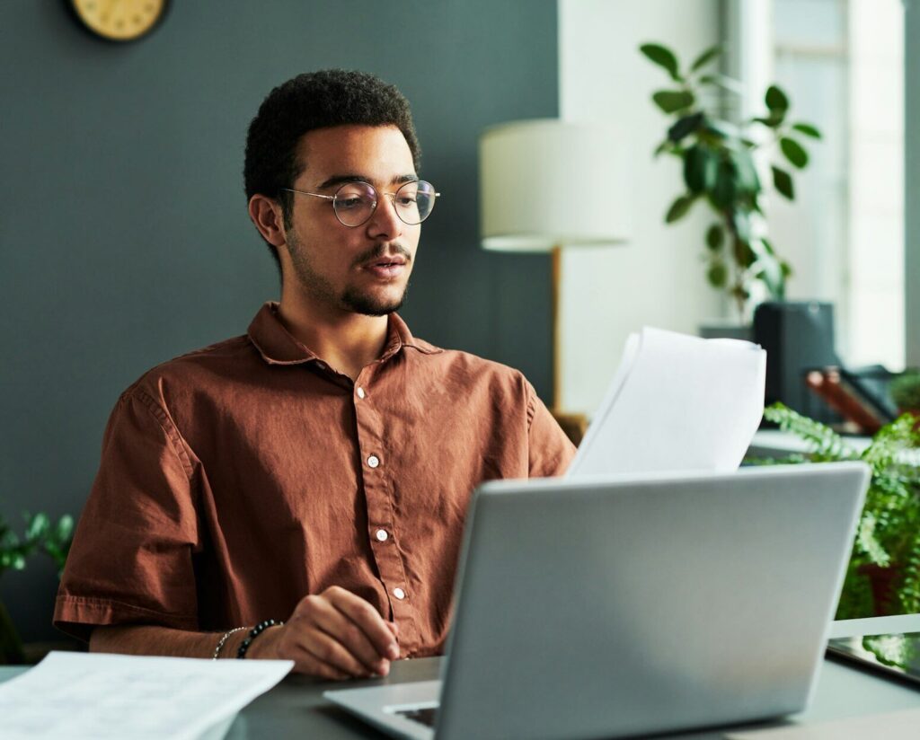 Young confident man with papers sitting in front of laptop